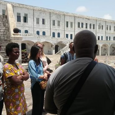A guide talking with visitors outside a historic building in Ghana